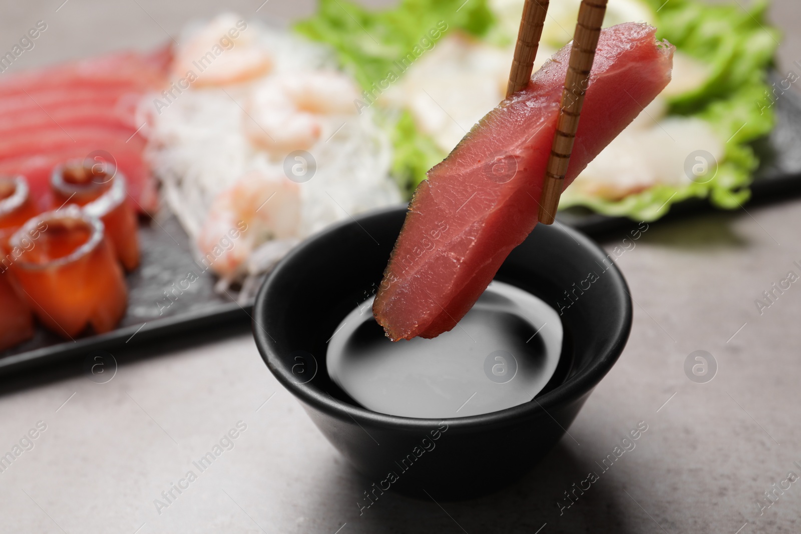 Photo of Chopsticks with raw tuna slice under soy sauce at grey table, closeup. Sashimi set