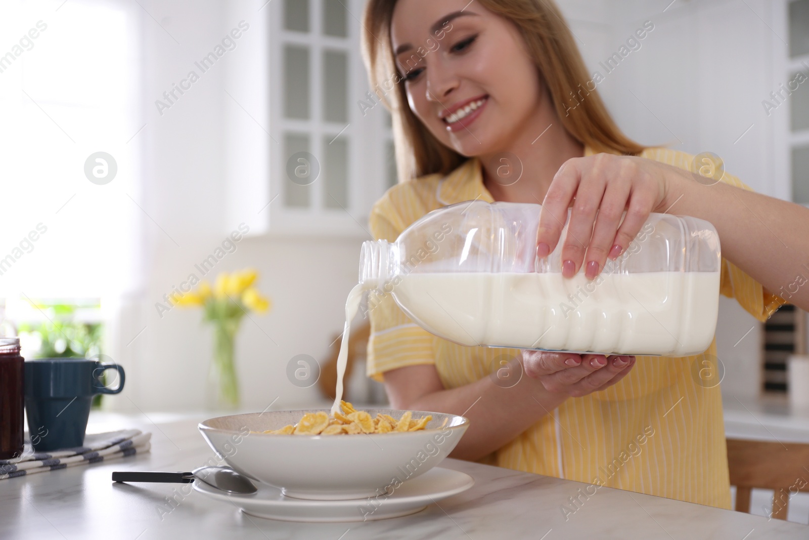 Photo of Young woman pouring milk from gallon bottle into plate with breakfast cereal at white marble table in kitchen