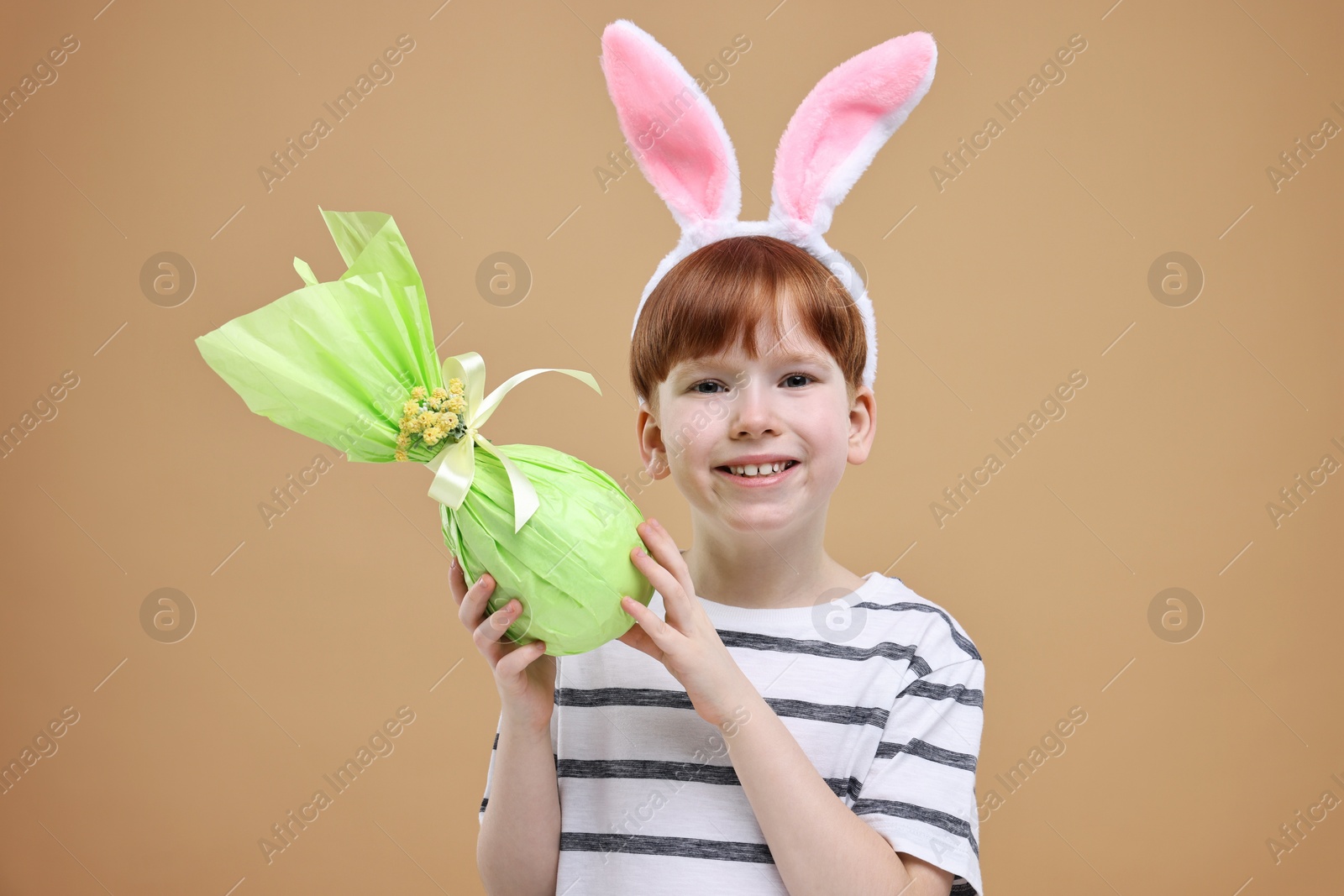 Photo of Easter celebration. Cute little boy with bunny ears and wrapped egg on dark beige background