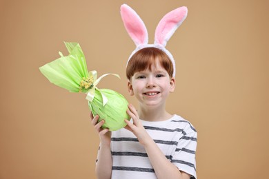 Easter celebration. Cute little boy with bunny ears and wrapped egg on dark beige background