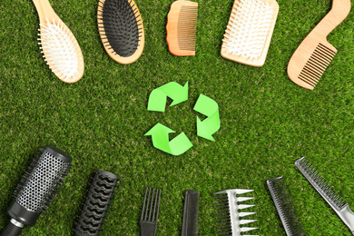 Recycling symbol, plastic and wooden hairbrushes on green grass, flat lay