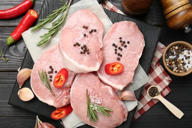 Photo of Pieces of raw pork meat, chili peppers and spices on black wooden table, flat lay