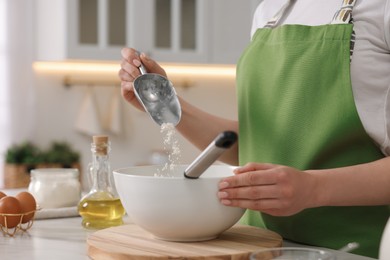 Photo of Making bread. Woman putting flour into bowl at white table in kitchen, closeup