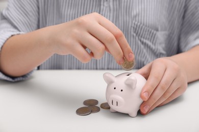 Woman putting coin into ceramic piggy bank at white wooden table, closeup. Financial savings
