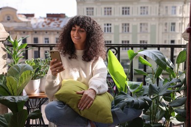 Photo of Beautiful young woman using smartphone surrounded by houseplants on balcony