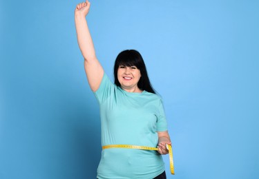 Happy overweight mature woman measuring waist with tape on light blue background