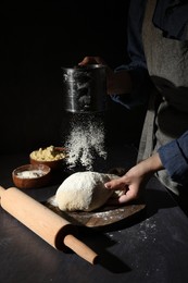 Photo of Woman sprinkling flour over dough at black table, closeup