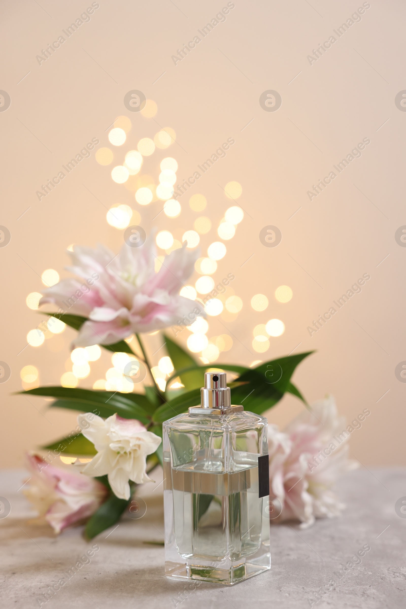 Photo of Bottle of perfume and beautiful lily flowers on table against beige background with blurred lights, space for text