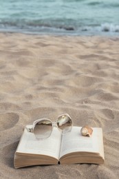 Photo of Beautiful sunglasses, book and shell on sand near sea, space for text