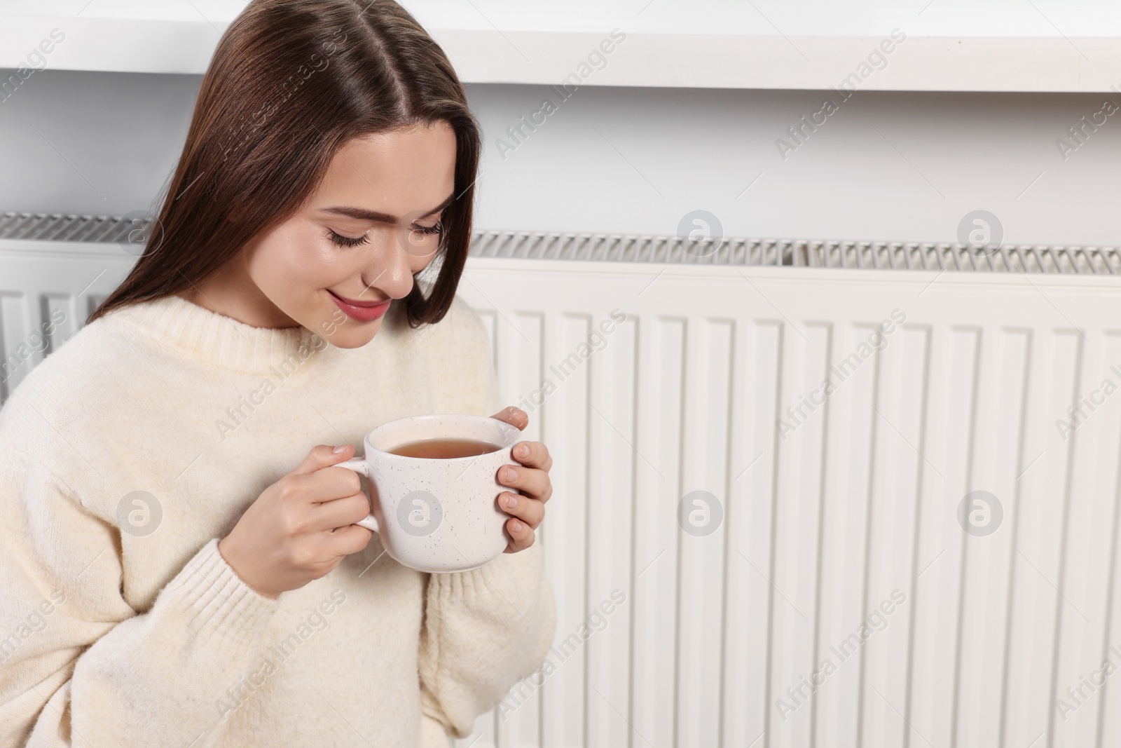 Photo of Woman holding cup with hot drink near heating radiator indoors