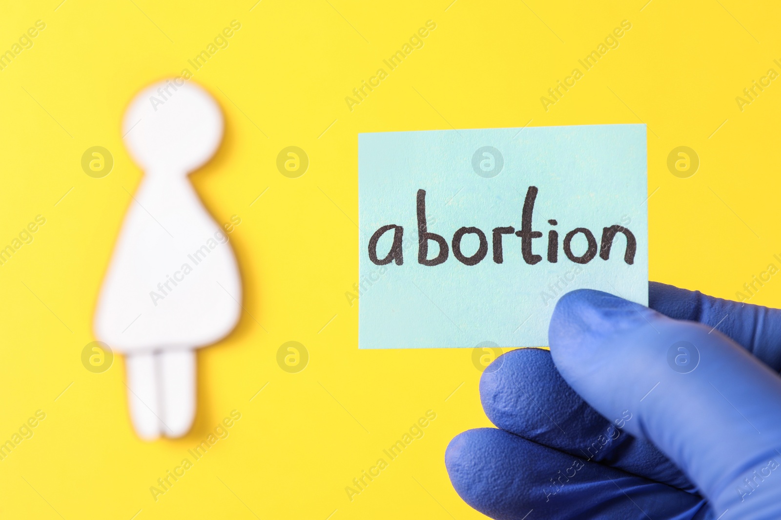 Photo of Doctor holding card with word ABORTION and paper cutout of pregnant woman on yellow background, closeup. Space for text