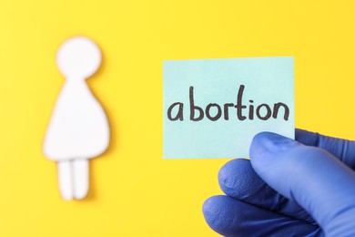 Photo of Doctor holding card with word ABORTION and paper cutout of pregnant woman on yellow background, closeup. Space for text