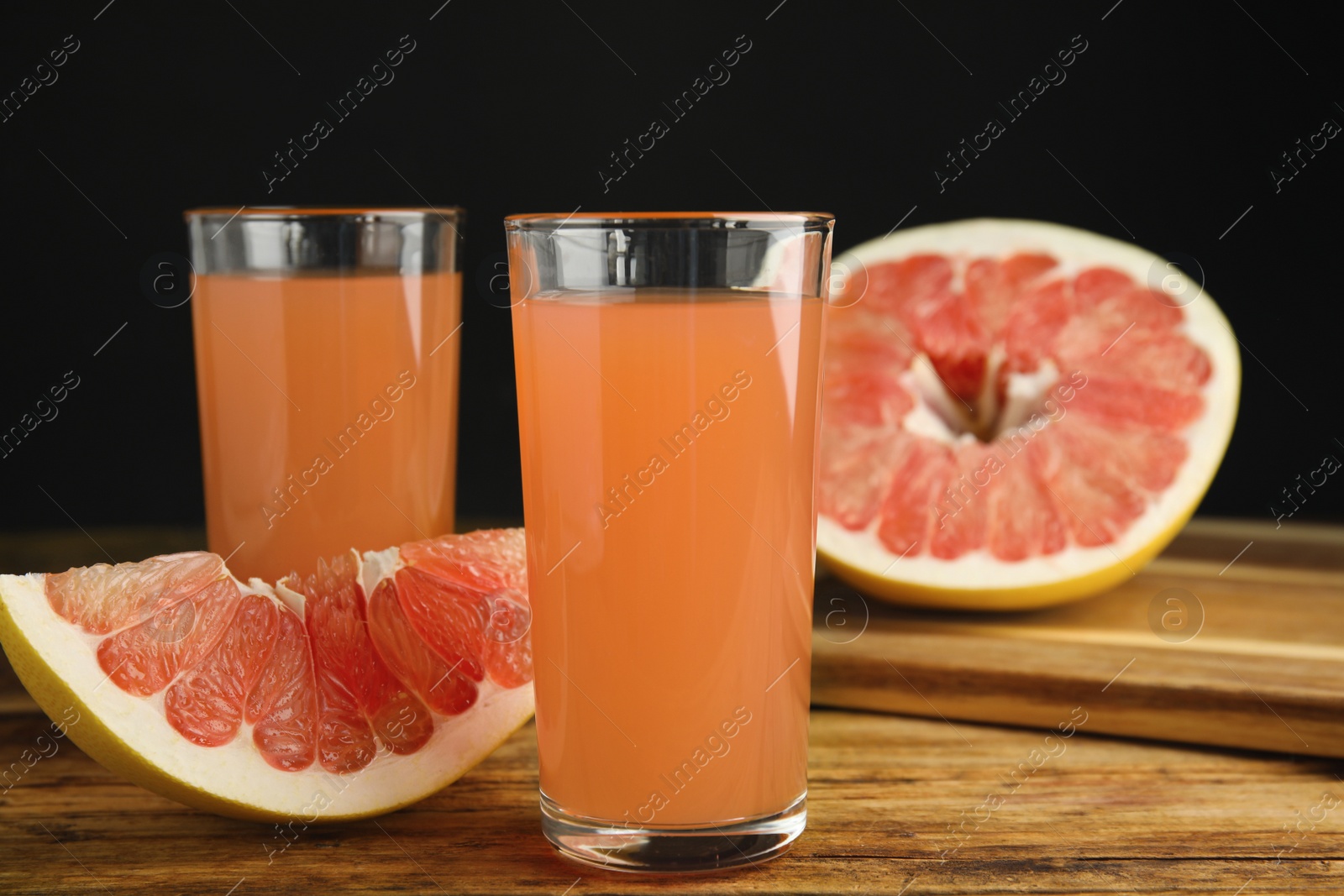 Photo of Glasses of pink pomelo juice and fruit on wooden table against black background