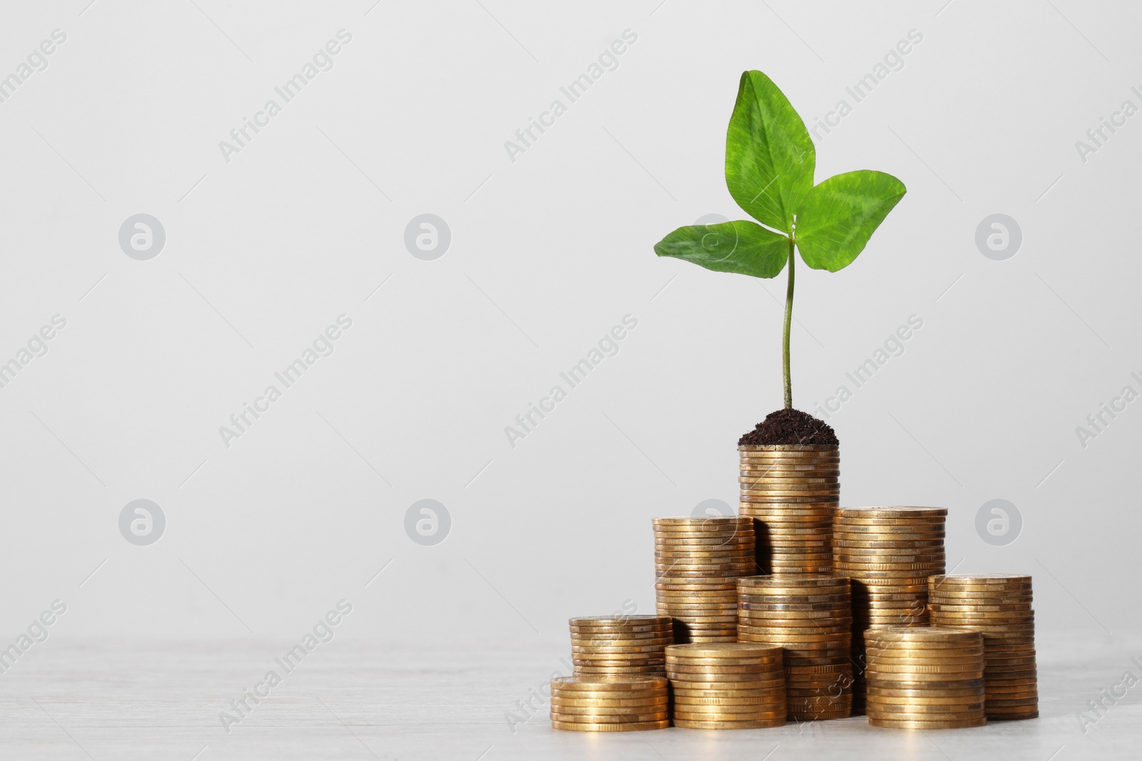 Photo of Stacks of coins with green sprout on white table against light grey background, space for text. Investment concept