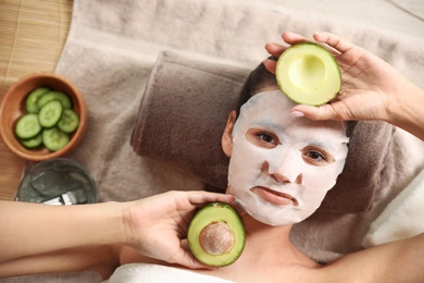 Photo of Beautiful woman with cotton mask sheet on face and cut avocado relaxing in spa salon, top view