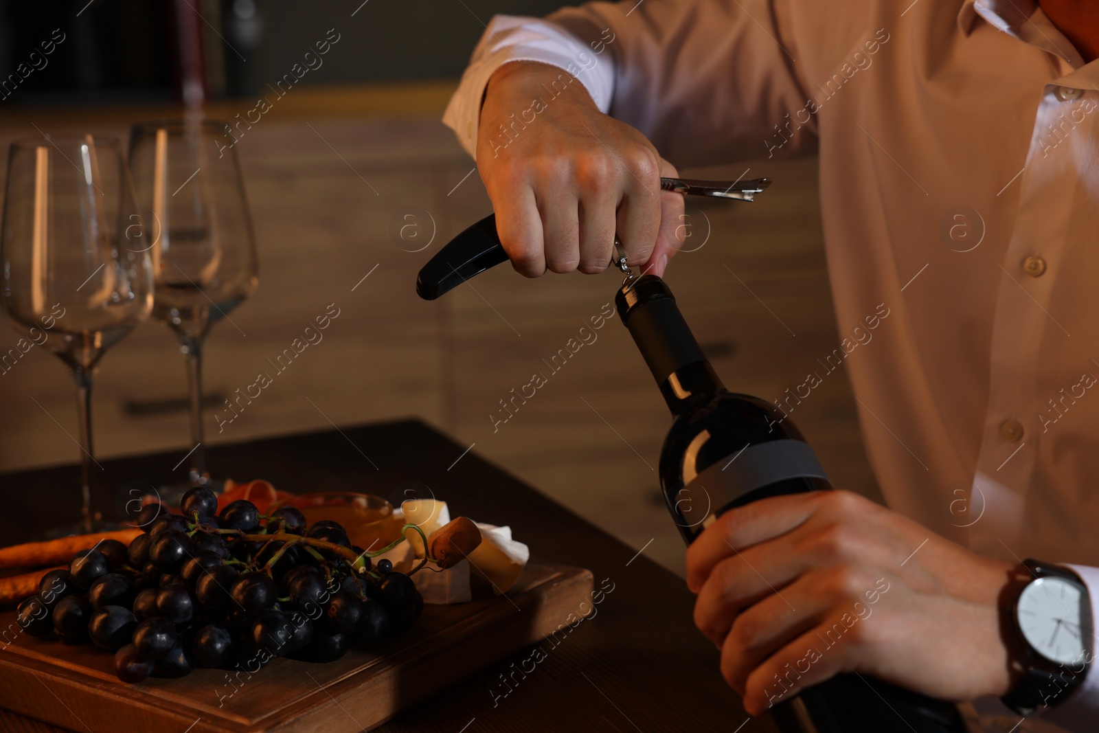 Photo of Romantic dinner. Man opening wine bottle with corkscrew at table indoors, closeup