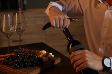 Romantic dinner. Man opening wine bottle with corkscrew at table indoors, closeup