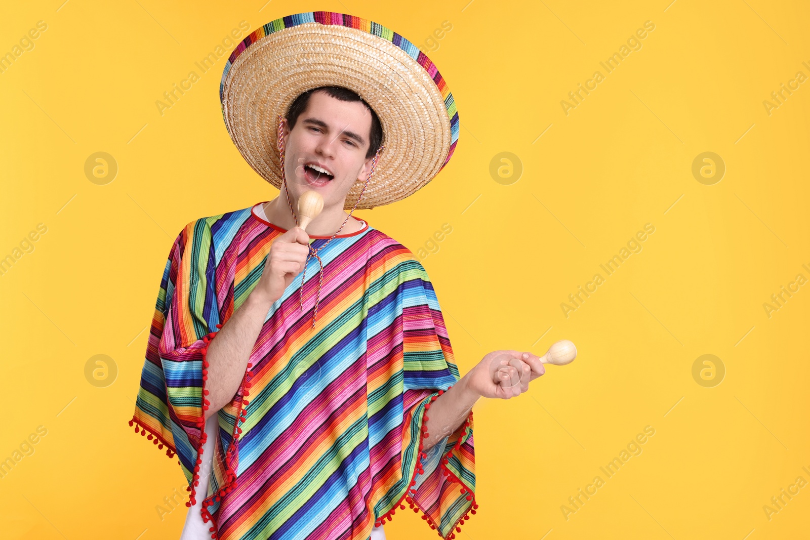 Photo of Young man in Mexican sombrero hat and poncho with maracas on yellow background. Space for text