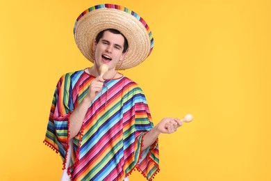 Young man in Mexican sombrero hat and poncho with maracas on yellow background. Space for text