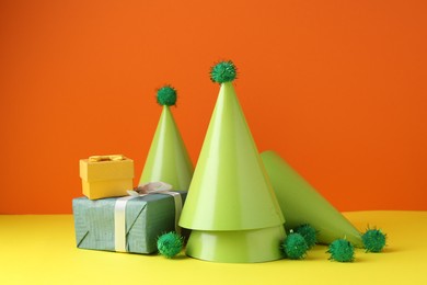 Party hats and gift boxes on yellow table against orange background