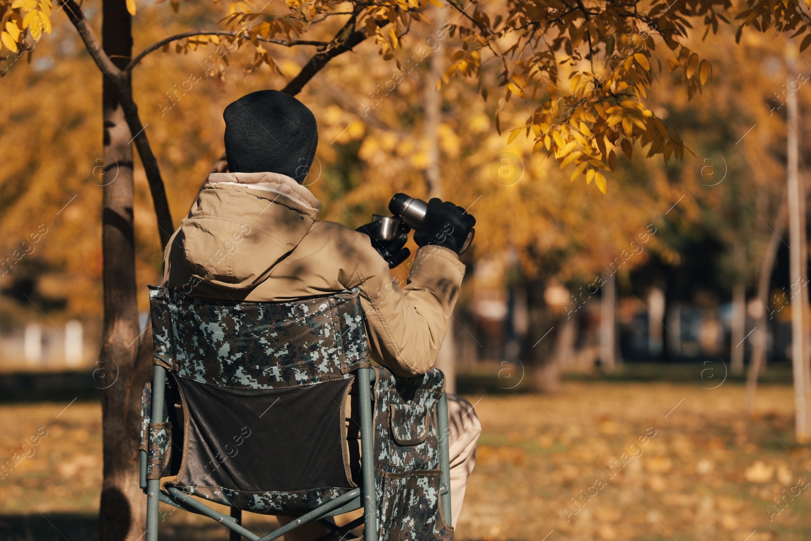 Photo of Man with thermos sitting in camping chair outdoors on autumn sunny day, back view