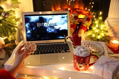 MYKOLAIV, UKRAINE - DECEMBER 23, 2020: Woman with cocoa watching Harry Potter movie on laptop near fireplace indoors, closeup. Cozy winter holidays atmosphere