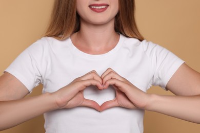 Young woman making heart with hands on beige background, closeup. Volunteer concept