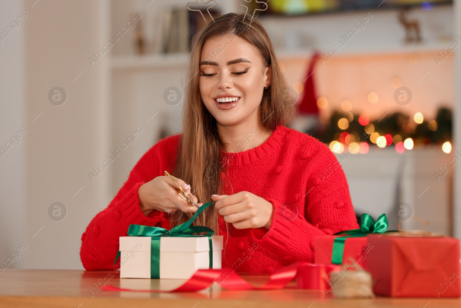 Photo of Beautiful young woman cutting ribbon. Decorating Christmas gift at table indoors