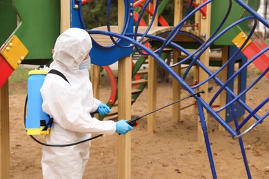 Photo of Woman wearing chemical protective suit with disinfectant sprayer on playground. Preventive measure during coronavirus pandemic