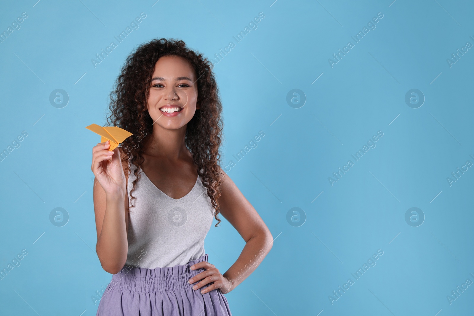 Photo of Beautiful African-American woman playing with paper plane on light blue background. Space for text