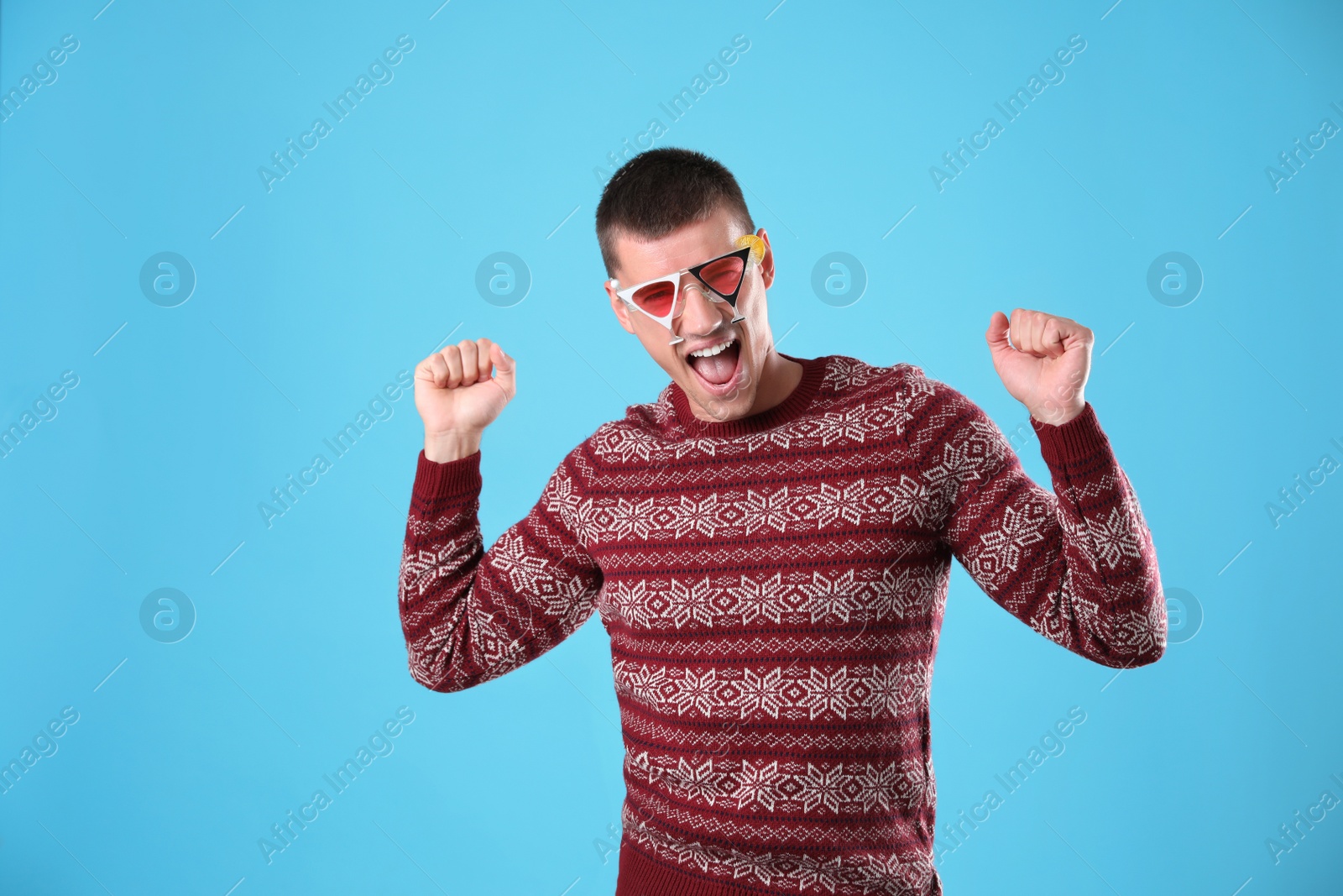 Photo of Emotional young man in Christmas sweater and party glasses on light blue background