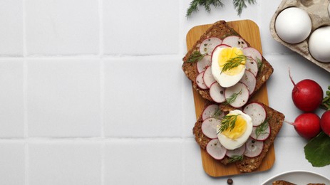 Photo of Tasty sandwiches with boiled egg, radish and ingredients on white tiled table, flat lay. Space for text