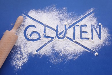Photo of Rolling pin and crossed out word Gluten written with flour on blue background, flat lay