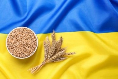 Photo of Bowl of wheat grains and ears on national Ukrainian flag, flat lay. Space for text