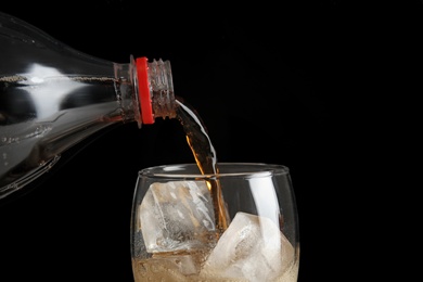 Photo of Pouring refreshing cola from bottle into glass with ice cubes on black background, closeup