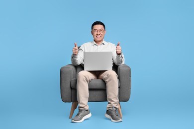 Photo of Happy man with laptop showing thumb up gesture while sitting in armchair on light blue background