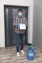 Photo of Courier in face mask with clipboard and bottles of cooler water in entryway. Delivery during coronavirus quarantine