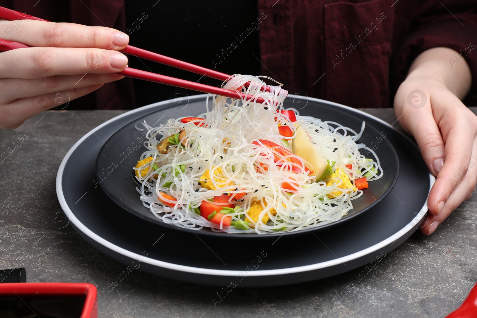 Photo of Stir-fry. Woman with chopsticks eating tasty rice noodles with meat and vegetables at grey textured table, closeup