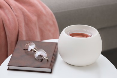 Photo of Wax air freshener, book and glasses on white table indoors. Interior element