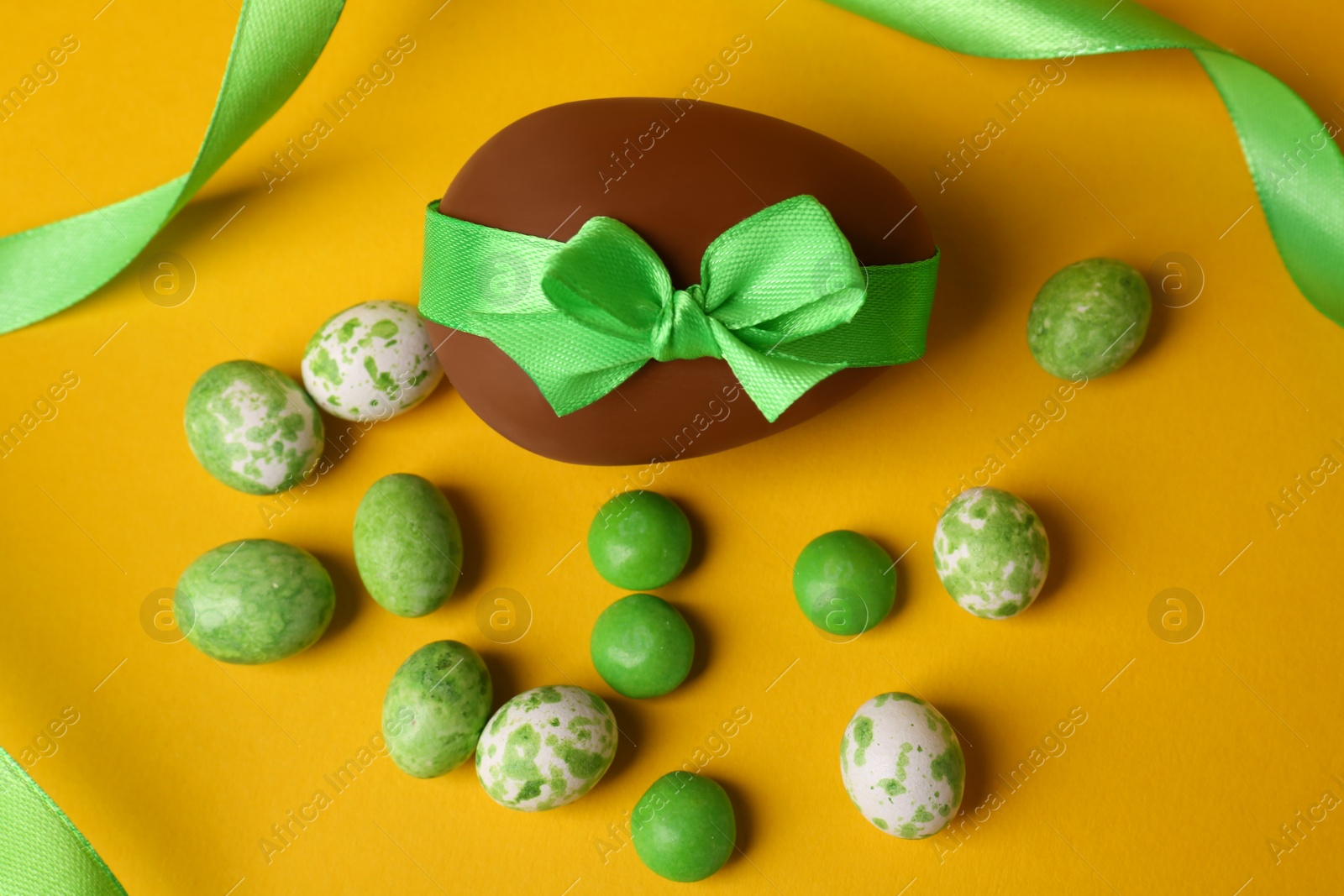 Photo of Tasty chocolate egg with green ribbon and candies on yellow background, flat lay
