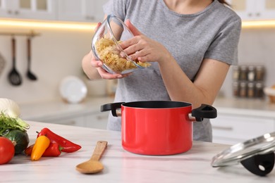 Photo of Housewife adding raw pasta in pot at white marble table in kitchen, closeup. Cooking process