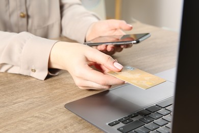 Online payment. Woman using smartphone and credit card near laptop at wooden table indoors, closeup