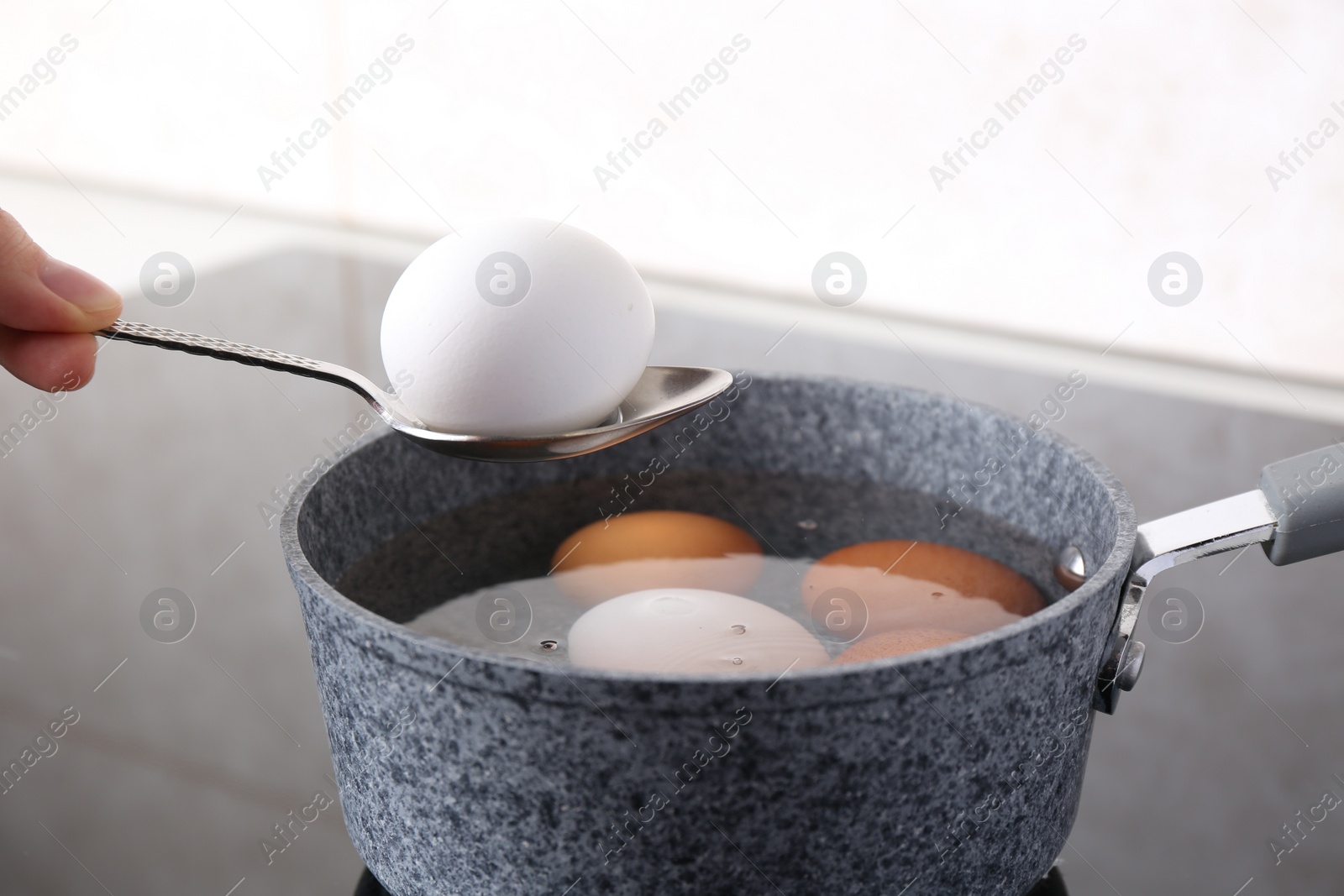 Photo of Woman holding spoon with boiled egg above saucepan on electric stove, closeup