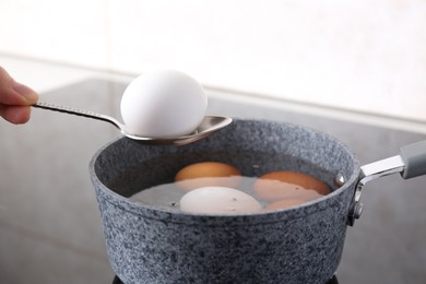 Woman holding spoon with boiled egg above saucepan on electric stove, closeup