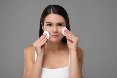 Young woman cleaning her face with cotton pads on grey background