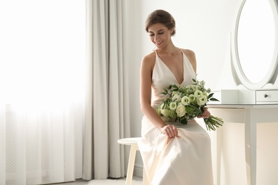 Photo of Young bride in wedding dress with beautiful bouquet near mirror indoors