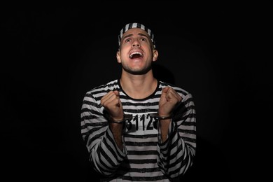 Photo of Emotional prisoner in striped uniform with handcuffs on black background