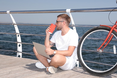 Attractive man with laptop and bike near sea on sunny day