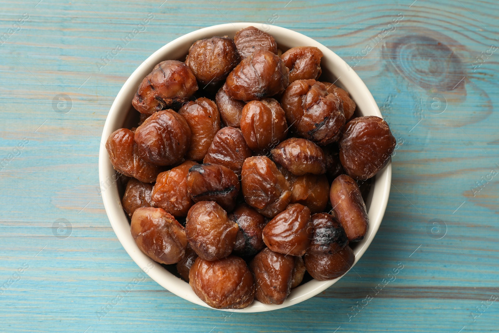 Photo of Roasted edible sweet chestnuts in bowl on light blue wooden table, top view