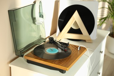 Stylish turntable with vinyl disc and headphones on white chest of drawers at home